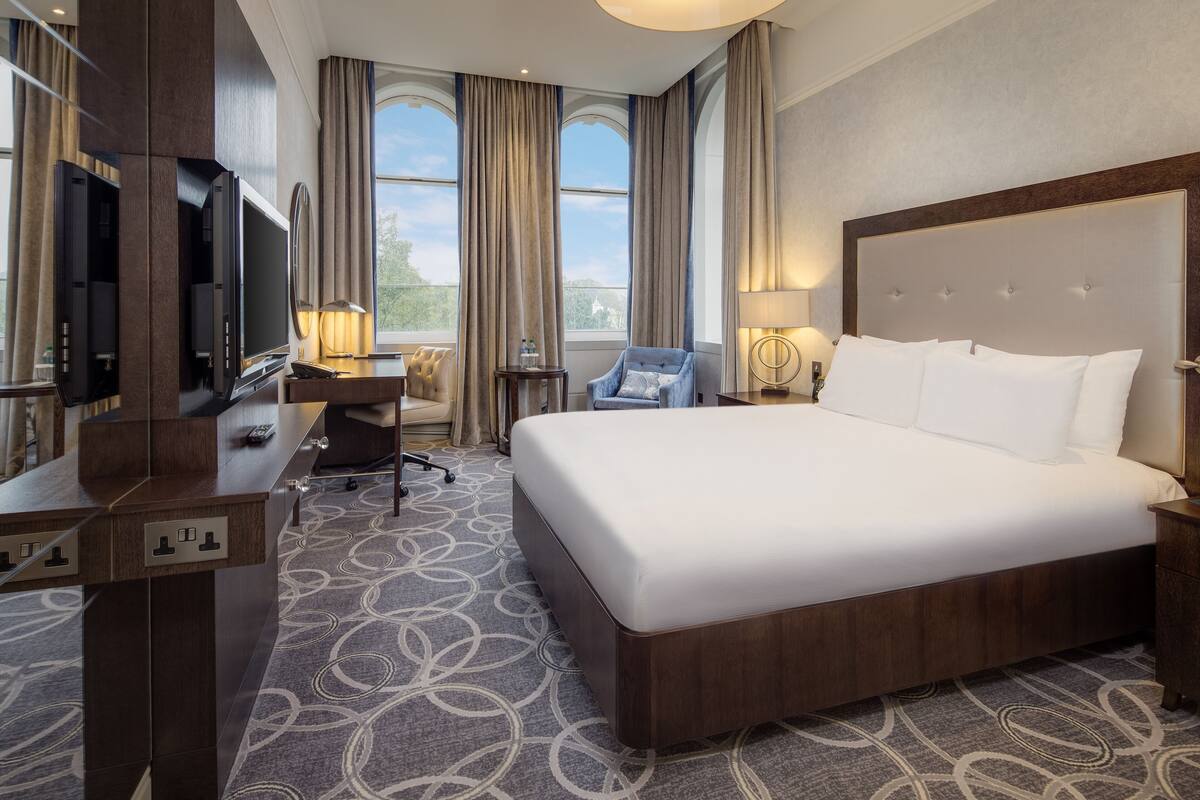 Ein Hotelzimmer mit einem großen Bett, einem Schreibtisch mit Stuhl, einem Fernseher und einem Fenster mit Vorhängen.