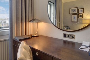 A hotel room with a wooden desk, a leather chair, a round mirror, and a window with curtains.