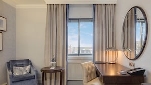 A hotel room with a bed, a desk, a chair, a round table, a lamp, a mirror, and a view of the city through the window.
