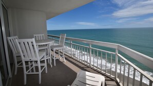 A balcony with white chairs and a table, overlooking a beach with waves and a clear blue sky. - 1001 Waters Edge Resort 3 Bedroom Condo by RedAwning (Murrells Inlet)