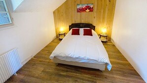 A bedroom with a wooden sloped ceiling, a bed with white bedding and red pillows, two bedside lamps, and a picture on the wall. - Gîte Lilas Sensation (Thury)