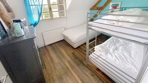 A dormitory room with bunk beds, a window with blue curtains, a radiator, and a wooden desk with a lamp. - Gîte Lilas Sensation (Thury)
