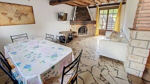 A rustic room with a stone floor, a fireplace, a television mounted on the wall, and a table with chairs. - Gîte Lilas Sensation (Thury)