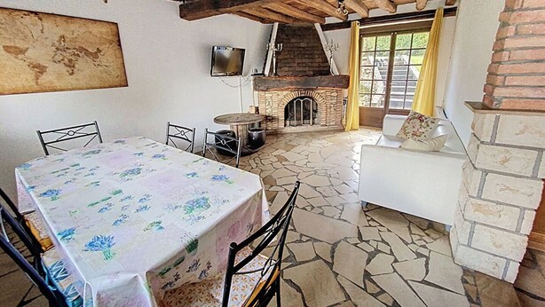 A rustic room with a stone floor, a fireplace, a television mounted on the wall, and a table with chairs. - Gîte Lilas Sensation (Thury)