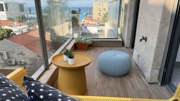 Un balcón con una silla amarilla, un taburete redondo y dos macetas.