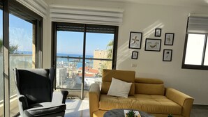 Una sala de estar moderna con un sofá, un sillón y una mesa de centro, ventanas amplias con vista a la ciudad y varias obras de arte enmarcadas en la pared.