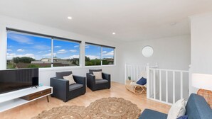 A spacious living room with a large TV, two dark armchairs, a wicker chair, a round mirror, and a view of the neighborhood through large windows. - Marlin Beach House - South Beach (Port Fairy)