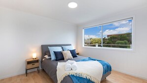 A bedroom with a bed, a nightstand, a window with a view of buildings and trees, and a ceiling light. - Marlin Beach House - South Beach (Port Fairy)