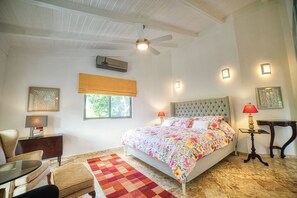 Un dormitorio con cama, ventilador de techo, ventana con persiana, mesita de noche con lámpara, silla y una pequeña mesa.