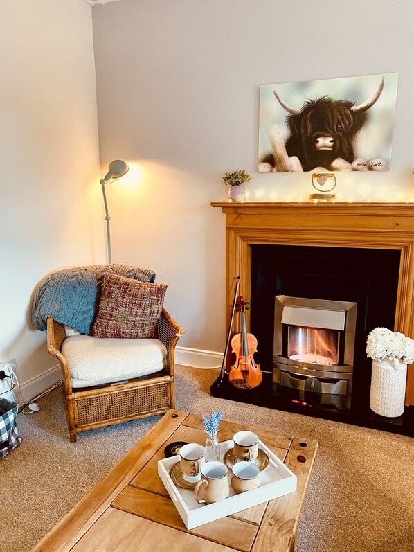 A cozy living room with a fireplace, a sofa, a coffee table with a tray of cups, and a violin leaning against the wall.