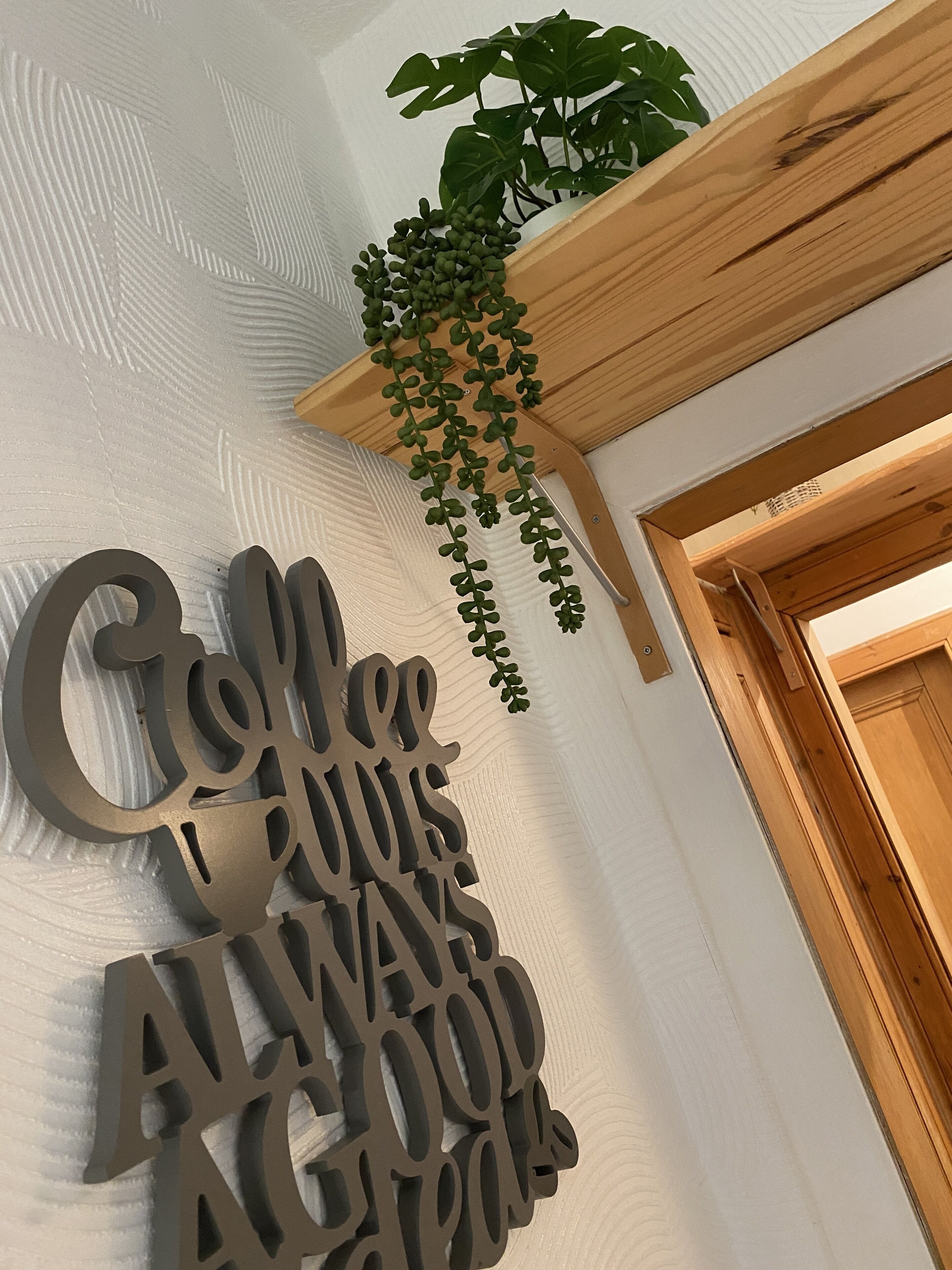 Un mur orné d’une enseigne métallique décorative sur laquelle on peut lire : « La bonne nourriture est toujours synonyme de bonne compagnie. » Une étagère en bois, sur laquelle est suspendue une plante, se trouve au-dessus de l’enseigne.