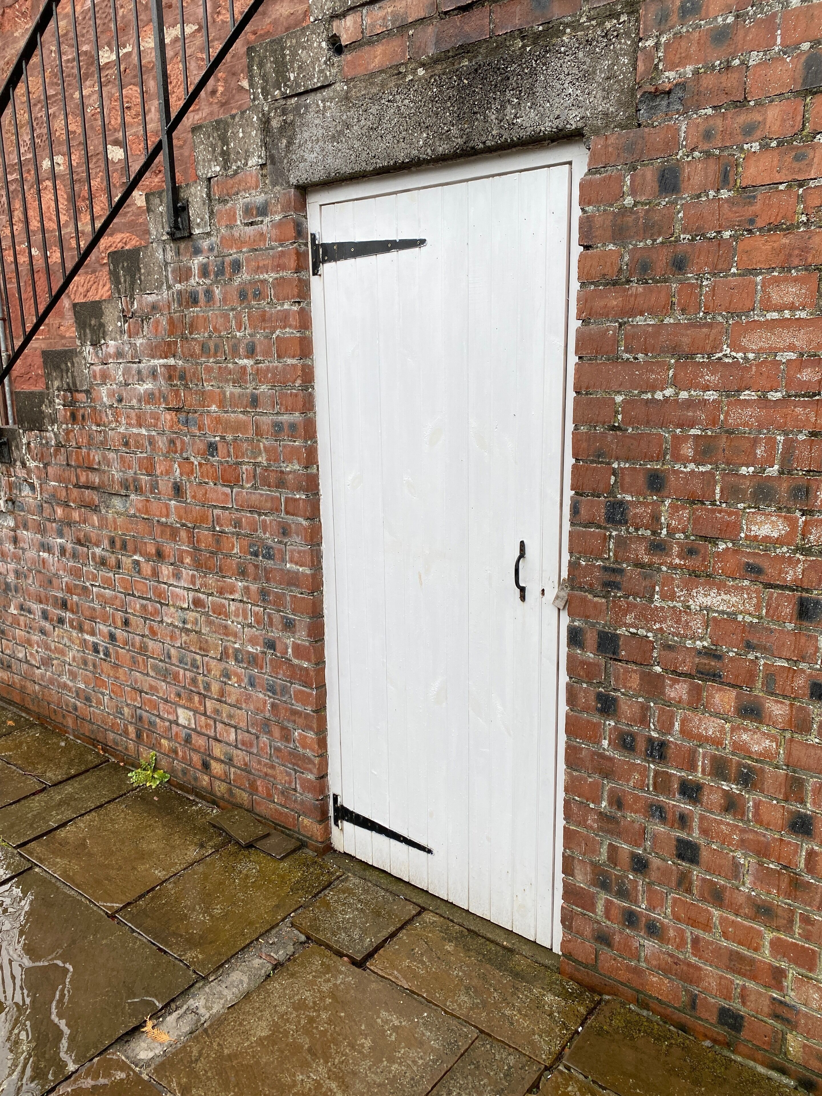 A white door with a black handle and a metal latch, set in a brick wall with a metal staircase to the left.