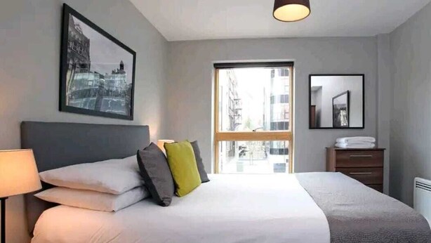 A modern hotel room with a large bed, a bedside table with a lamp, a window with a view of buildings, and a framed picture on the wall. - 2 Bed Apartment Sleeps 4 With Free Parking (Salford)