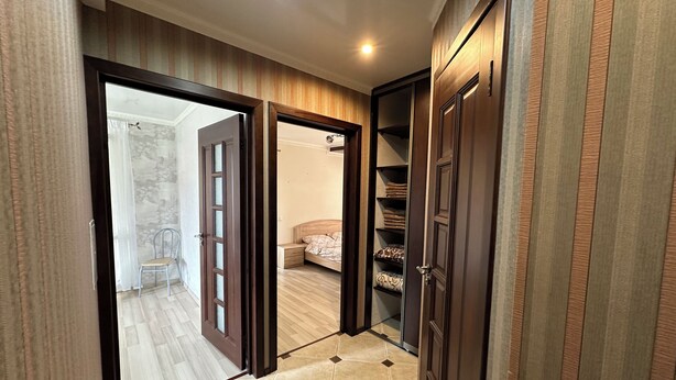 A hallway with a wooden door leading to a bedroom with a bed and a nightstand. There is a built-in shelf unit with folded clothes. -  PaulMarie Apartment on Podgorodskaya (Brest)