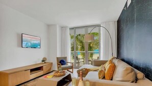 A modern hotel room with a large window, a flat-screen TV, a sofa, a chair, and a lamp.