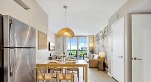 A modern kitchen with a stainless steel refrigerator, a dining table with chairs, a hanging light fixture, and a view of the outdoors through large windows. - 2BR w/ Balcony + Amazing Pool + Gym (Hallandale Beach)