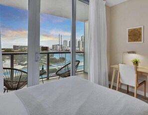 A hotel room with a bed, a desk, a chair, and a view of a cityscape and water. - Spectacular Apartment w/Balcony+ Pool+ Near Beach (Hallandale Beach)