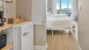 A hotel room with a bed, a minibar, a small desk with a coffee maker, and a view of the city through large windows. - Spectacular Apartment w/Balcony+ Pool+ Near Beach (Hallandale Beach)