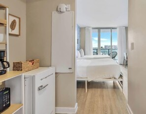 A hotel room with a bed, a minibar, a small desk with a coffee maker, and a view of the city through large windows. - Spectacular Apartment w/Balcony+ Pool+ Near Beach (Hallandale Beach)