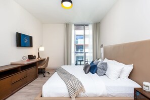 Une chambre d’hôtel avec un lit, un bureau, une chaise, une lampe et une vue sur les bâtiments par la fenêtre.