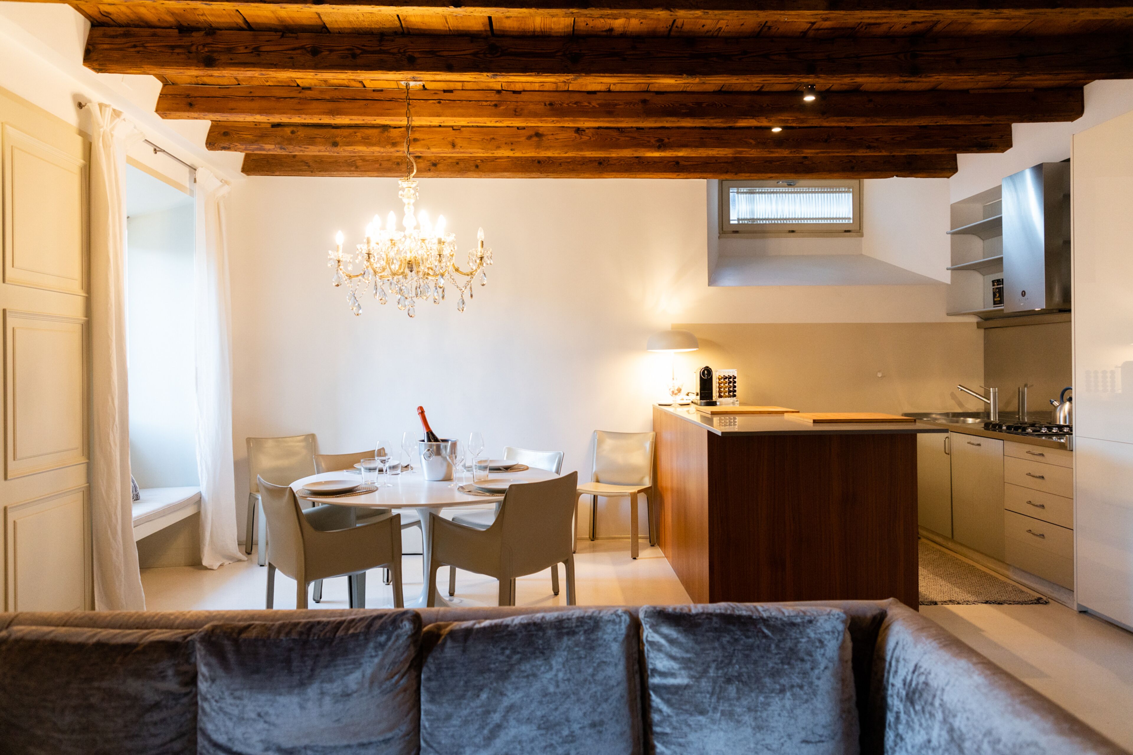 A modern kitchen with a wooden ceiling, a dining area with a chandelier, and a sofa in the foreground.