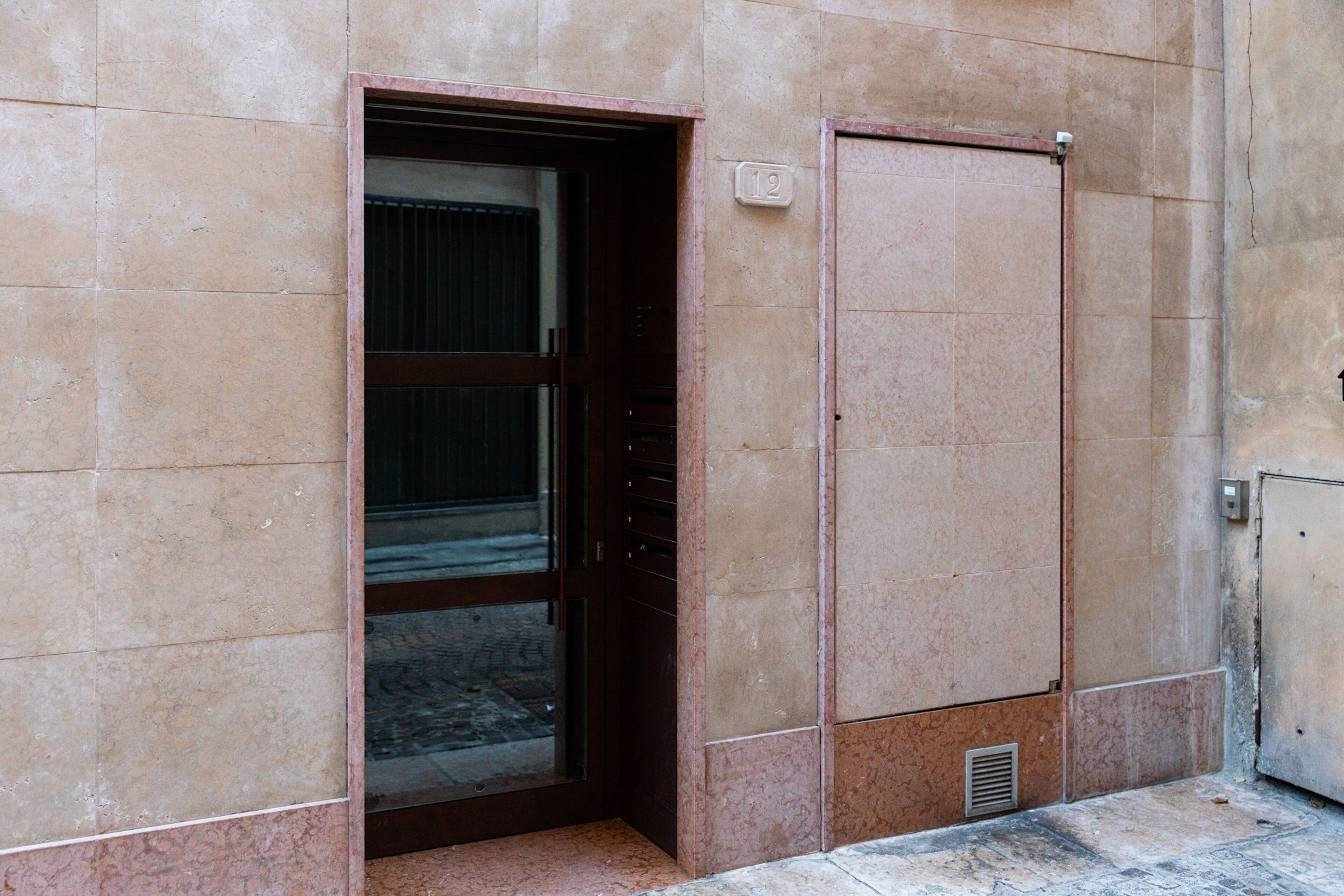 Ingresso di un edificio con una porta contrassegnata dal numero '12', una griglia di ventilazione sottostante e un muro rivestito di piastrelle.