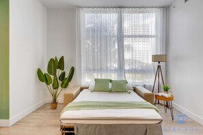 Un dormitorio con una cama, una mesita de noche con lámpara, una planta en maceta y una ventana con cortinas translúcidas.
