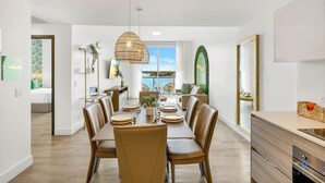 A modern dining area with a large table set for six, surrounded by leather chairs. A hanging light fixture is above the table, and a large window provides a view of the outdoors.