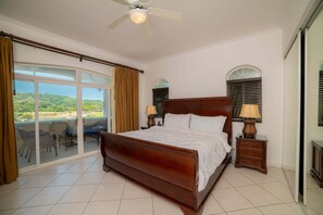 A bedroom with a large bed, a ceiling fan, a nightstand with a lamp, and a view of a balcony.