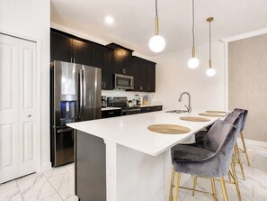 Une cuisine moderne dotée d’un îlot central, d’appareils électroménagers en acier inoxydable, de meubles de rangement aux couleurs sombres et de luminaires suspendus.