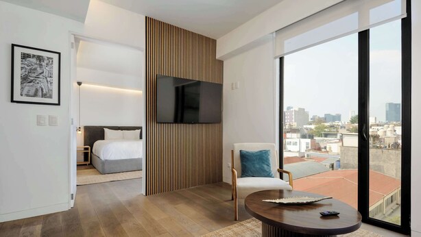 A modern hotel room with a large TV, a bed, a chair, and a balcony with a city view. - w* | Modern 1BR in New Building in Roma Norte (Ciudad de México)