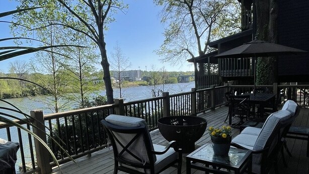A wooden deck with outdoor furniture, a fire pit, and a view of a lake and buildings in the distance. - Beautiful 4 Bedroom River Front House in Downtown (Augusta)