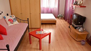 Een knus, klein kamertje met een bed, een bank, een rode tafel, een televisie en een kledingkast.