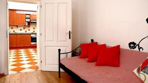 Een slaapkamer met een bed, rode kussens, een witte deur en uitzicht op een keuken met houten kasten en een geruit vloertje.
