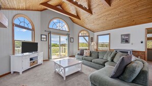 A spacious living room with a large sectional sofa, a white coffee table, and a television on a white cabinet. Large arched windows provide ample natural light and views of the outdoors. - OCEANSIDE:  Private Pool, beautiful home on full acre lot! (Corolla)