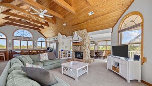 A spacious living area with a stone fireplace, a television, and a large window overlooking the outdoors. - OCEANSIDE:  Private Pool, beautiful home on full acre lot! (Corolla)