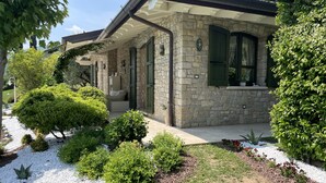 Una casa in pietra con un portico coperto, persiane verdi e un giardino curato con arbusti e fiori diversi.