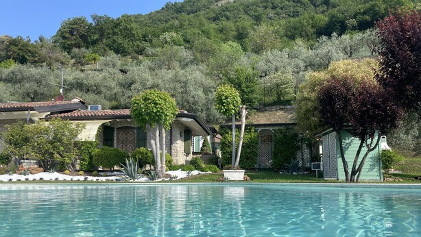 Una piscina con acqua cristallina, circondata da un giardino curato e un edificio in pietra con tetto in tegole.