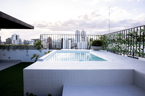 Une piscine sur le toit, offrant une vue panoramique sur la ville, entourée d’une clôture en métal et de verdure.