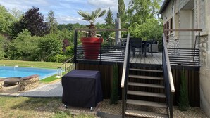 Una terraza de madera con mobiliario exterior, una parrilla y una piscina al fondo.