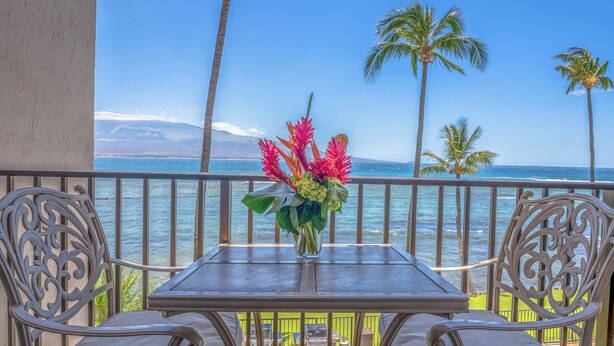 A balcony with a table and chairs, overlooking a beach with palm trees and the ocean. - *Lauloa 304* Maui Prime Oceanfront Condo, A/C! (Maalaea)