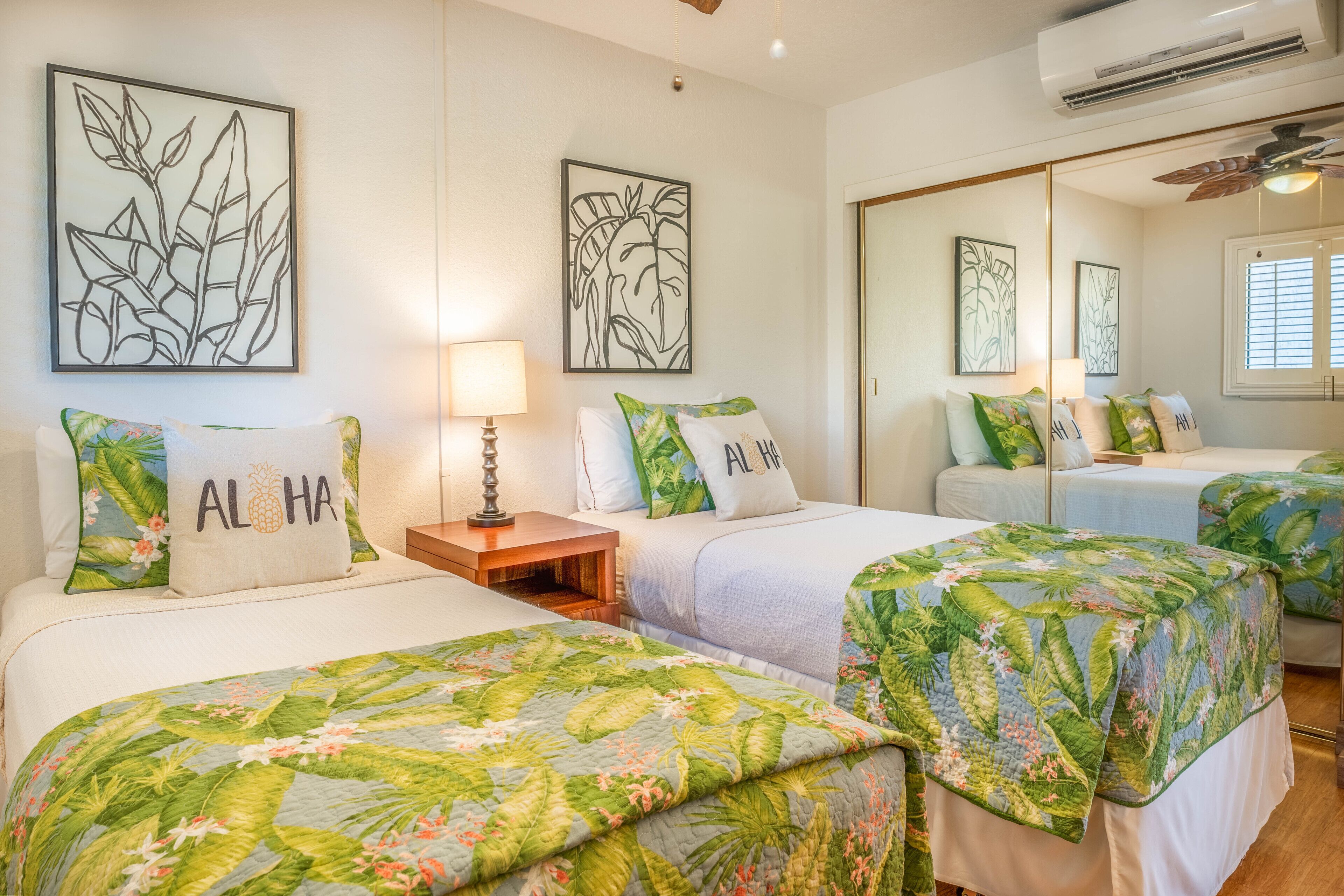 A hotel room with two beds, each with a floral bedspread and pillows with tropical prints. There is a nightstand with a lamp, and artwork depicting tropical leaves on the walls.