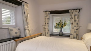 A bedroom with a bed, a window with curtains, a radiator, and a vase with flowers on the windowsill. - Green Stile Cottage (Windermere)