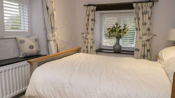 A bedroom with a bed, a window with curtains, a radiator, and a vase with flowers on the windowsill.