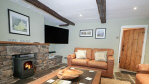 A cozy living room with a stone fireplace, a leather sofa, and a flat-screen TV mounted on the wall. - Green Stile Cottage (Windermere)