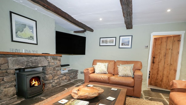 A cozy living room with a stone fireplace, a leather sofa, and a flat-screen TV mounted on the wall. - Green Stile Cottage (Windermere)