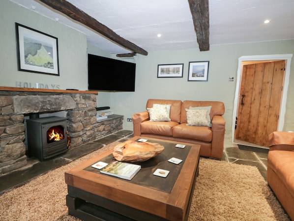 A cozy living room with a stone fireplace, a leather sofa, and a flat-screen TV mounted on the wall. - Green Stile Cottage (Windermere)