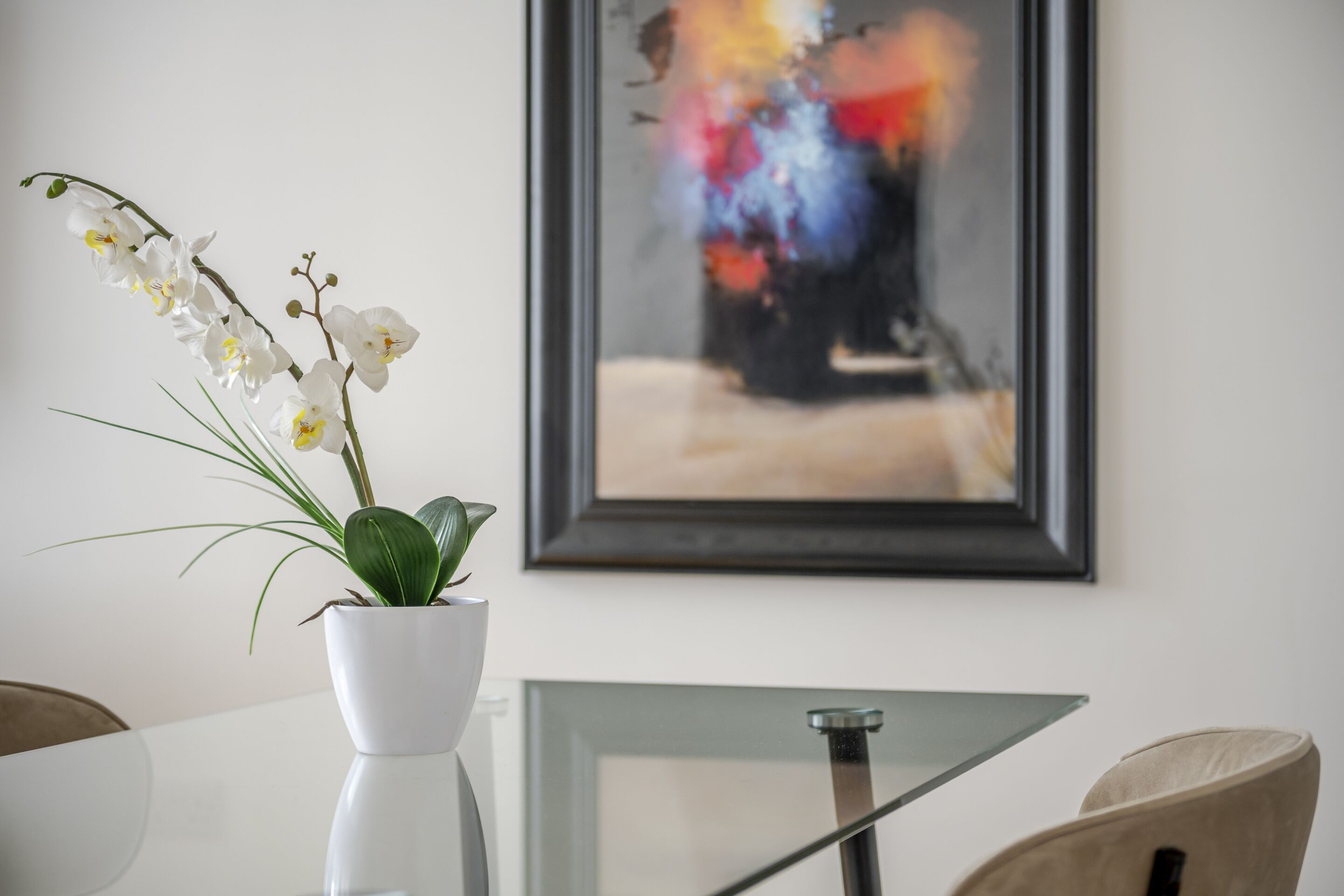 A white orchid in a white pot on a glass table with a framed abstract painting in the background.