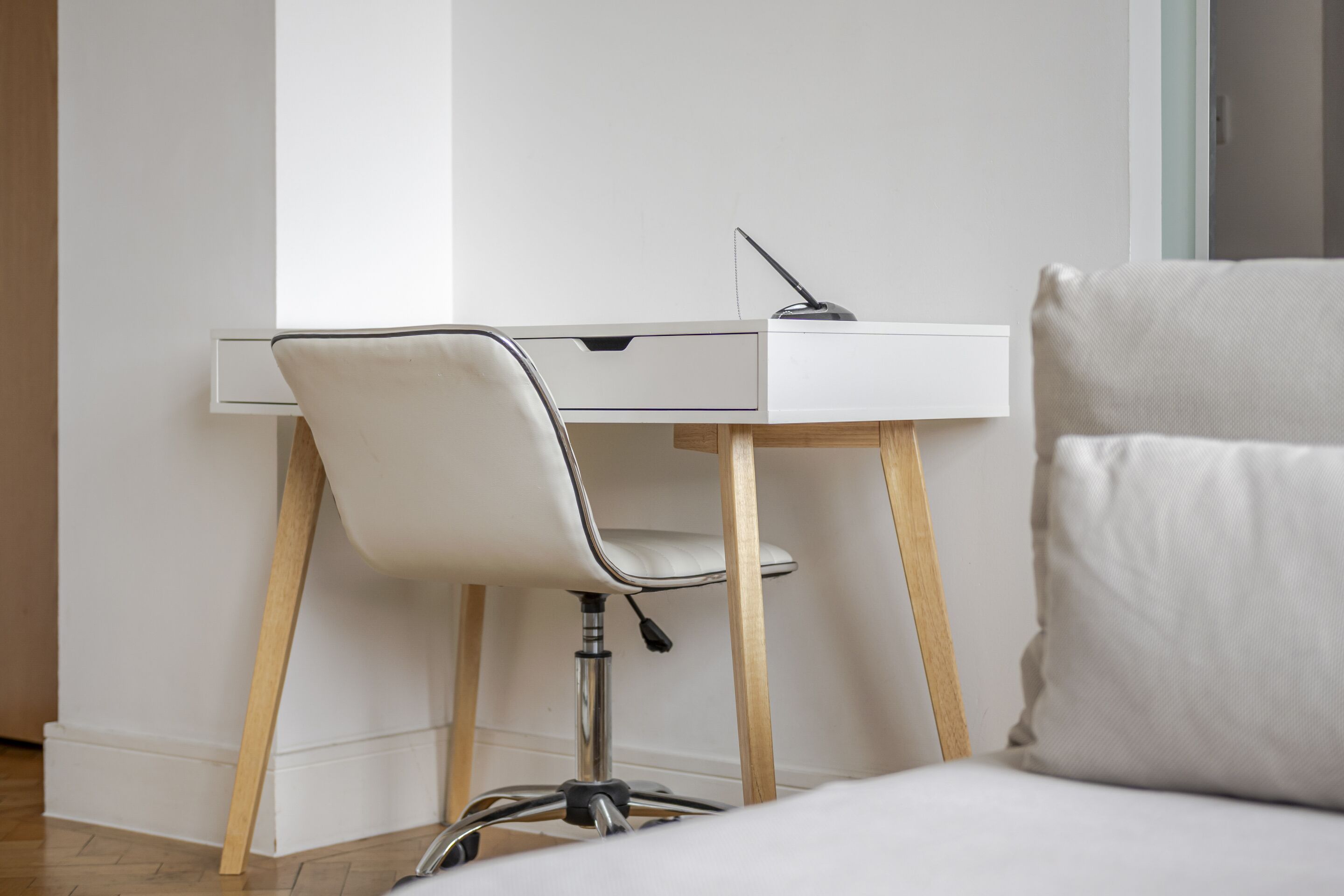 A modern office space with a white desk, a white chair, and a bed with a white pillow.