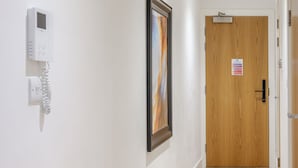 Un pasillo con un cuadro abstracto enmarcado, un detector de incendios y una puerta de madera con un cartel de emergencia.
